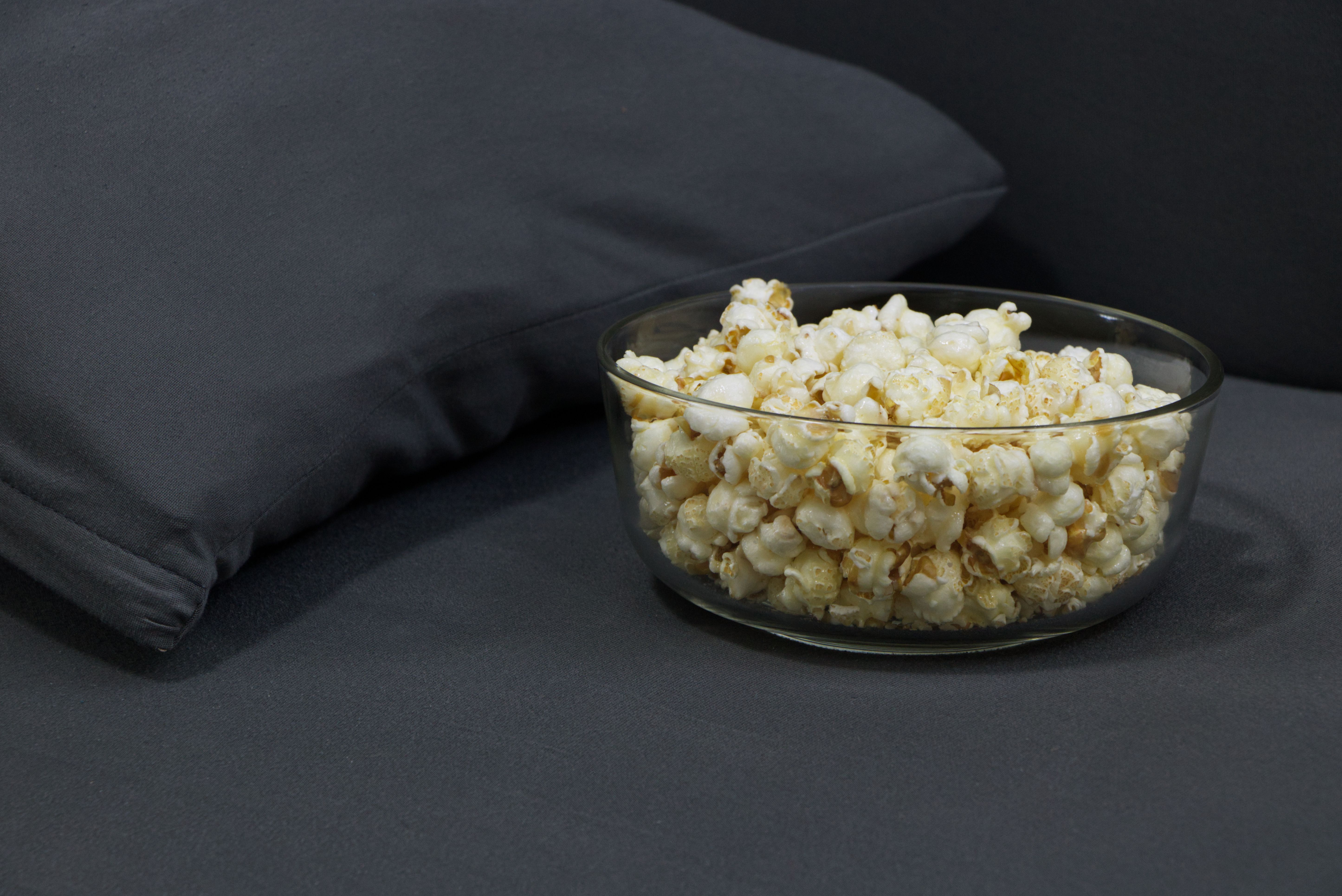 A bowl of popcorn on a dark sofa beside a remote control, captured in a photorealistic cozy movie-night scene